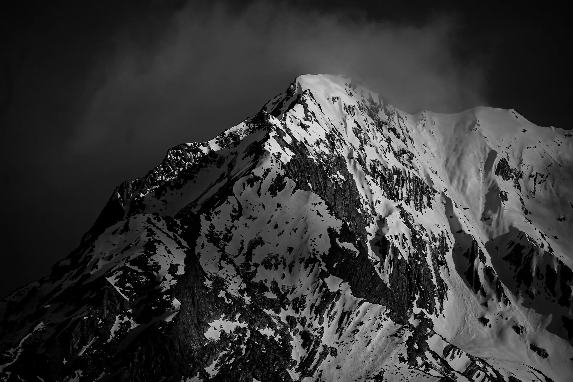 A snow-covered peak lit against a bank of dark cloud, in black and white.